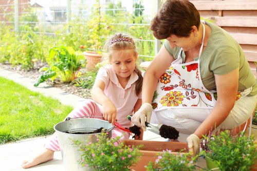 A woman and a young girl are working together in a neatly maintained garden that features a vibrant flower bed with various blooming plants, bordered by a low brick or stone edge. They are planting or tending to flowers in a rectangular wooden planter box placed on a paved patio area. The garden background includes lush green grass, potted plants, and a variety of shrubs and small trees, with a wire fence and garden structures visible in the distance. The woman is wearing an apron and gloves, carefully handling soil or plants, while the girl, dressed in a light-colored top and striped pink trousers, observes and assists. The scene is set outdoors on a bright, sunny day, highlighting the fresh greenery and colourful flowers, supporting professional gardening and landscaping services in Greenwich and surrounding areas. The overall environment emphasizes outdoor maintenance and garden care in a residential setting, with natural light enhancing the vibrant colours and textures of the plants and garden elements.