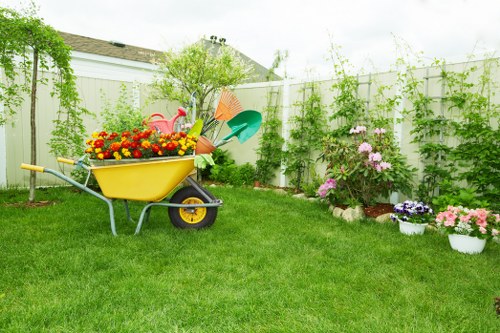 A well-maintained backyard garden featuring a lush, green lawn with evenly trimmed grass, bordered by vibrant flower beds containing pink, purple, and yellow flowers in bloom, some potted and others planted directly into the soil. A white wooden fence encloses the space, with climbing plants and greenery growing along its length, adding natural privacy. In the foreground, a yellow wheelbarrow filled with colourful garden decorations, such as a plastic flamingo and flower pots, is positioned on the grass. Gardening tools including a small blue shovel and an orange watering can are placed within or near the wheelbarrow for easy access. The background shows a mix of mature trees and shrubs, providing shade and structure to the outdoor environment, under an overcast sky with soft natural light. The scene showcases a tidy, inviting garden area suitable for lawn care and outdoor maintenance services offered by Landscapers Greenwich, emphasizing a peaceful, productive outdoor space in Greenwich postcodes.
