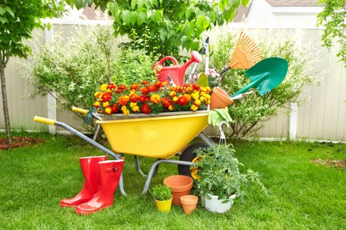 The image depicts a well-maintained backyard garden with a lush green lawn bordered by a white fence. In the center, there is a yellow wheelbarrow filled with vibrant red, orange, and yellow flowers, positioned on the grass. Surrounding the wheelbarrow are various gardening tools and accessories, including a red watering can, a green trowel, a blue garden fork, and a pair of bright red rubber boots placed on the ground. Several small plant pots and a white container with a green leafy plant are arranged nearby, indicating ongoing gardening activity. The background features a variety of leafy shrubs and trees, providing natural shade and a mature outdoor environment. The scene suggests a sunny day, ideal for garden maintenance, and highlights typical elements involved in garden care offered by landscaping professionals such as Landscapers Greenwich, serving areas like Greenwich and nearby postcodes.