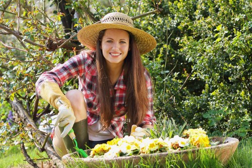 In a well-maintained garden, a young woman with long brown hair and a bright smile is kneeling beside a large, decorative flower bed filled with blooming yellow and white flowers. She is wearing a wide-brimmed straw hat, a plaid shirt with rolled-up sleeves, and gardening gloves, indicating she is engaged in outdoor gardening work. The garden features lush greenery with various shrubs and small trees in the background, suggesting a peaceful residential outdoor space in Greenwich. The flower bed is bordered with natural stone or wood, and the grass surrounding it appears dense and healthy, with a uniformly trimmed lawn area. Sunlight filters through the leaves, casting dappled light onto the scene, reflecting a clear, pleasant day suitable for gardening activities. This image highlights outdoor maintenance and landscaping efforts typical of local garden care services offered by companies like Landscapers Greenwich, with a focus on creating vibrant, well-organized garden spaces in the Greenwich area, postcode inferred from the pages related to gardening services in London.