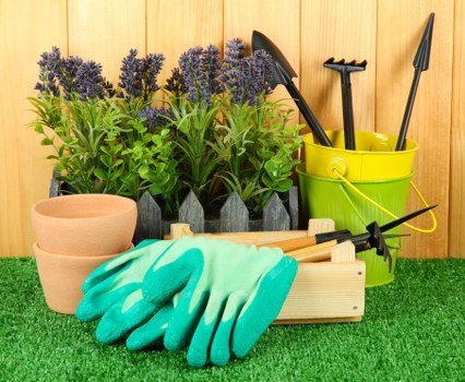 An organized gardening setup in an outdoor space shows several potted lavender plants with purple flowers and green foliage positioned at the back, with three unglazed terracotta pots atop a small wooden table in front. To the right, a bright yellow plastic bucket contains gardening tools such as trowels and a cultivator, with handles sticking out. A pair of light blue gardening gloves are laid on a wooden box filled with additional garden tools, and nearby, a green artificial grass surface covers the ground, enhancing the neat appearance of the space. The backdrop features wooden fencing, suggesting a backyard or front garden area designed for landscaping and outdoor maintenance. The scene captures tidy, practical elements typical of gardening services like pruning, planting, and general lawn care, aligning with the services offered by Landscapers Greenwich in the Greenwich area, within the postcode district.
