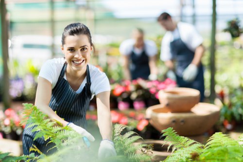 A smiling woman wearing a striped blue apron and white shirt is working in a brightly lit greenhouse or garden center, surrounded by lush green plants, including ferns and flowering plants in pots. She appears to be gardening or planting, with her hands engaged in tending to the greenery in front of her. In the background, two other individuals, also dressed in gardening attire, are slightly out of focus as they handle flowers or plants, indicating a collaborative gardening or landscaping activity. The environment features a glass structure allowing natural light to illuminate the vibrant, well-maintained plants, with shelves and display tables showcasing an array of colorful plants and garden decor. This setting emphasizes professional gardening and landscaping services in Greenwich, highlighting a dedicated outdoor space for planting and garden maintenance, with natural tones of green, brown, and hints of pink from flowers visible in the scene. Weather conditions suggest a warm, sunny day ideal for gardening work, supporting the idea of a professional gardening service being performed at this location.